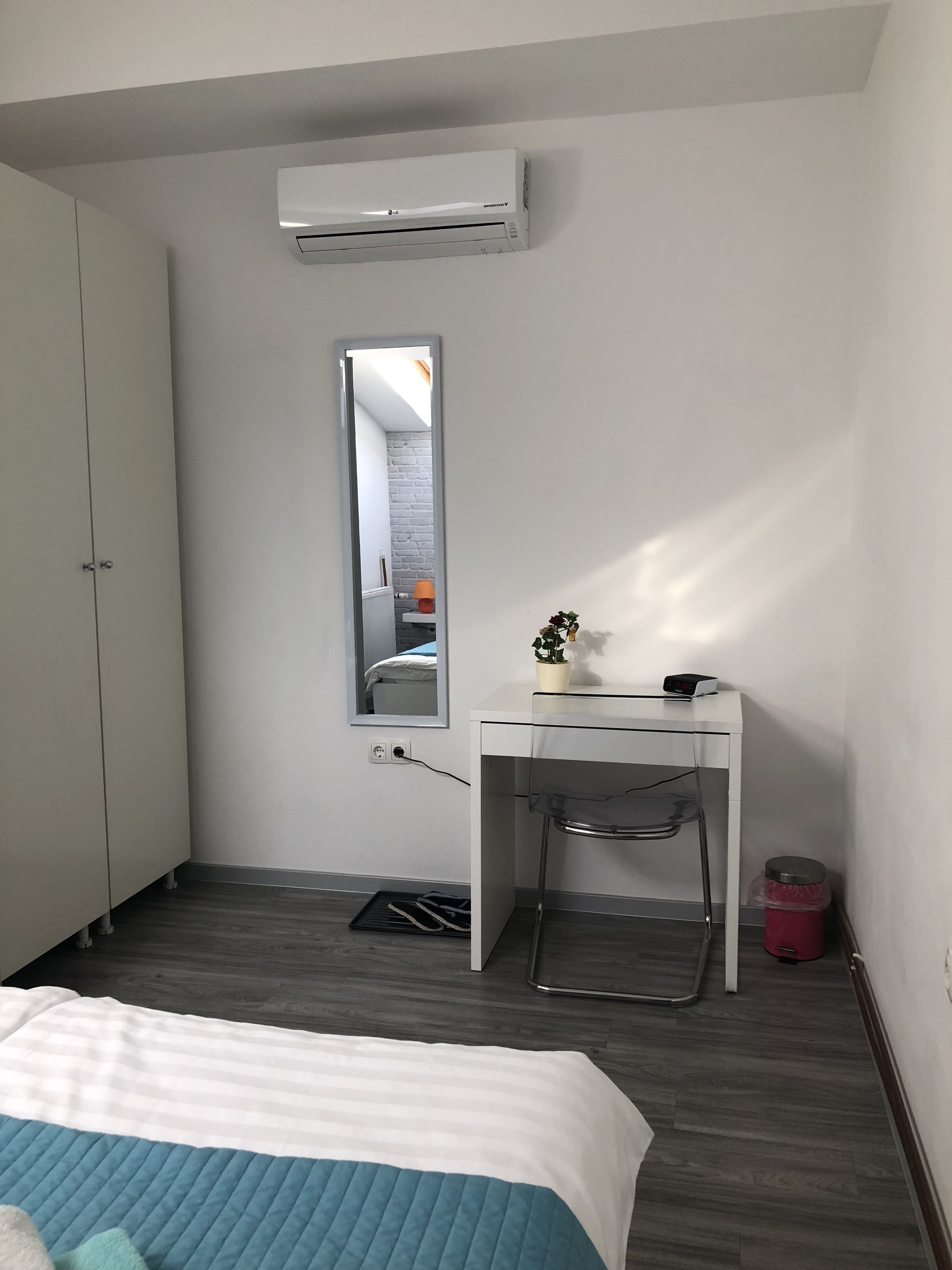 A bedroom with a bed, a desk, a chair, a mirror, an air conditioner, and a trash bin.
