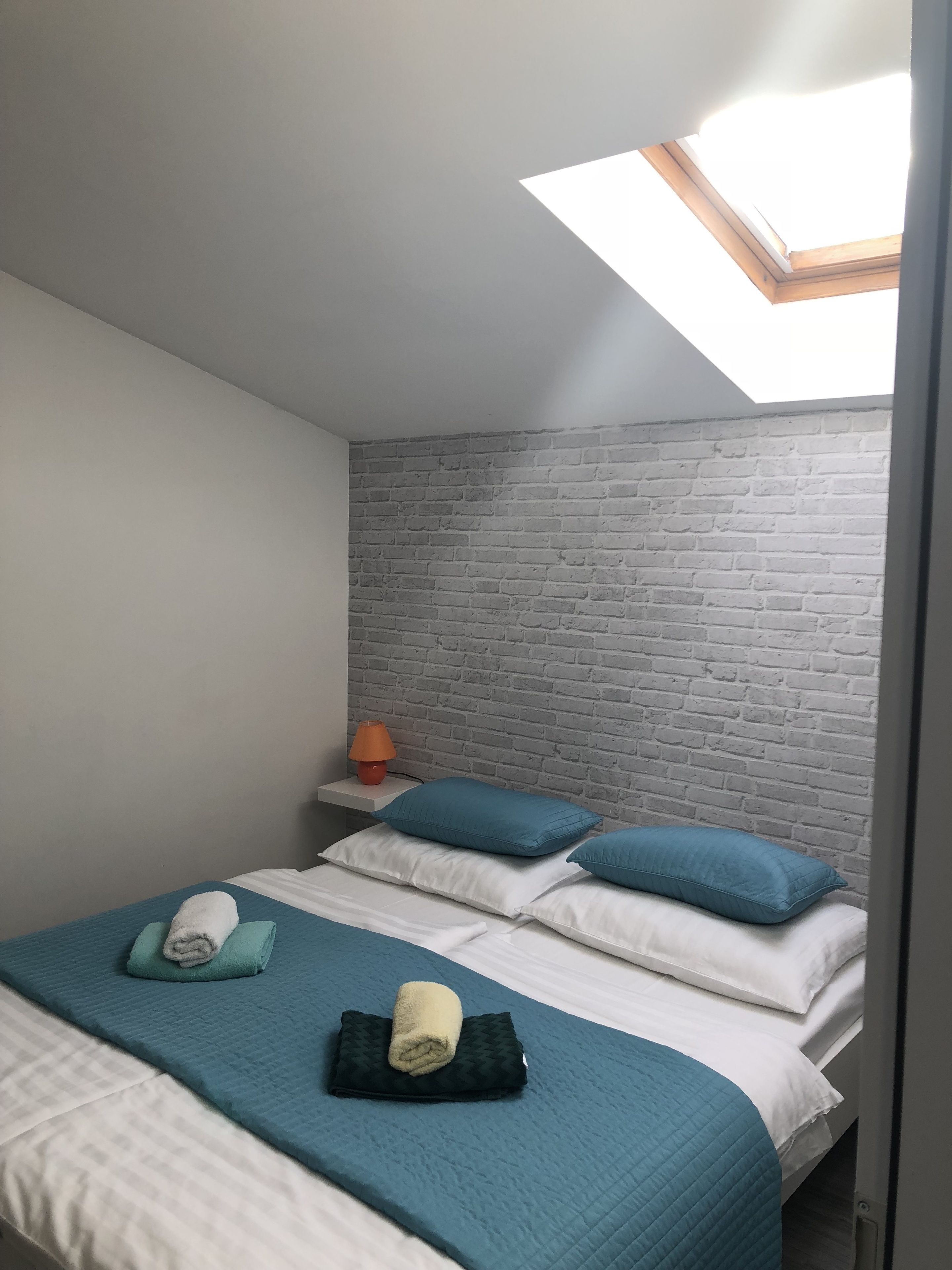 A room with a sloped ceiling, a skylight, a brick-patterned wall, a bed with blue and white bedding, and a nightstand with a lamp.