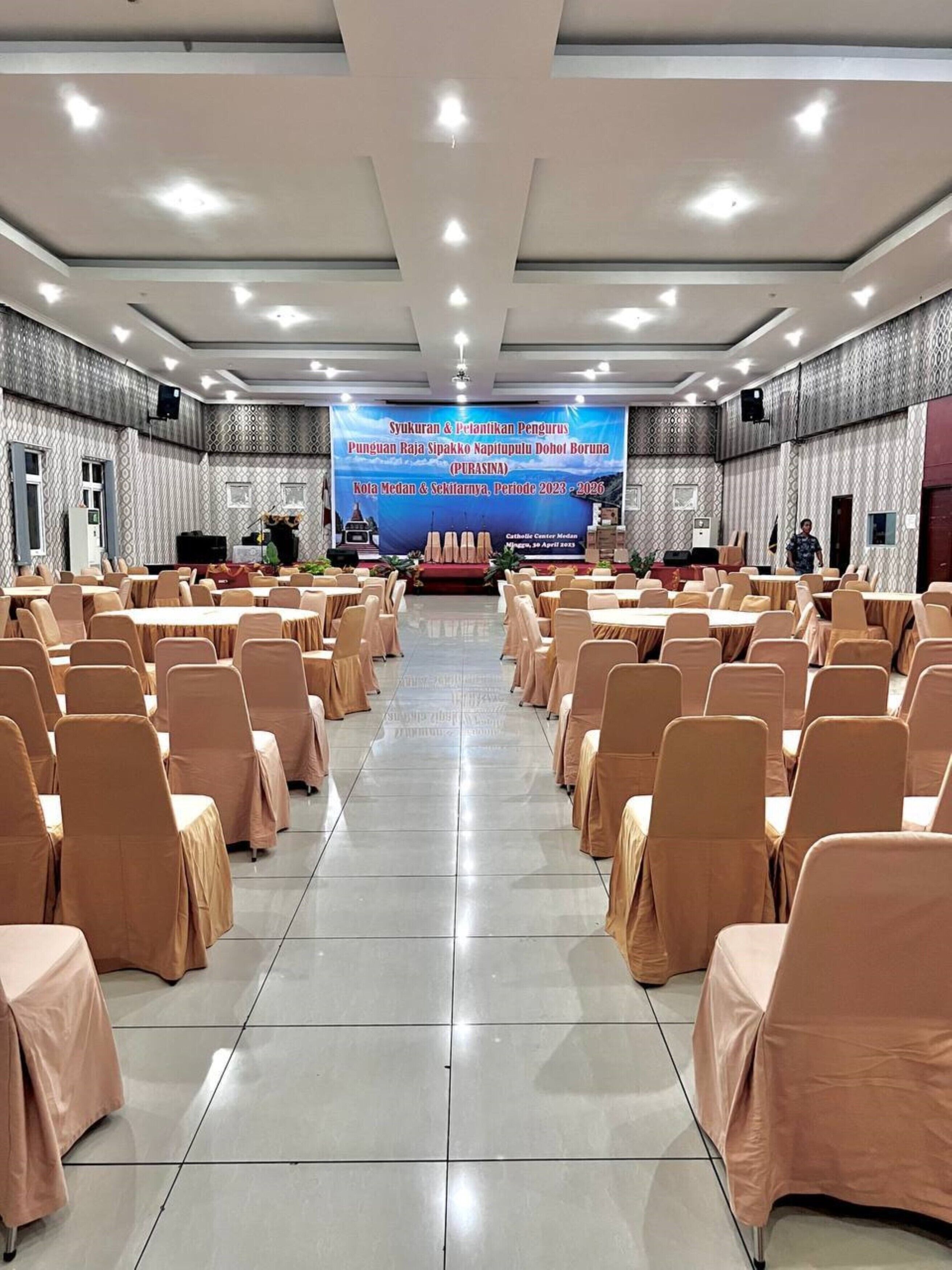 Une salle de conférence avec des rangées de tables et de chaises, une scène avec un grand écran à l’avant et des murs décorés de motifs.
