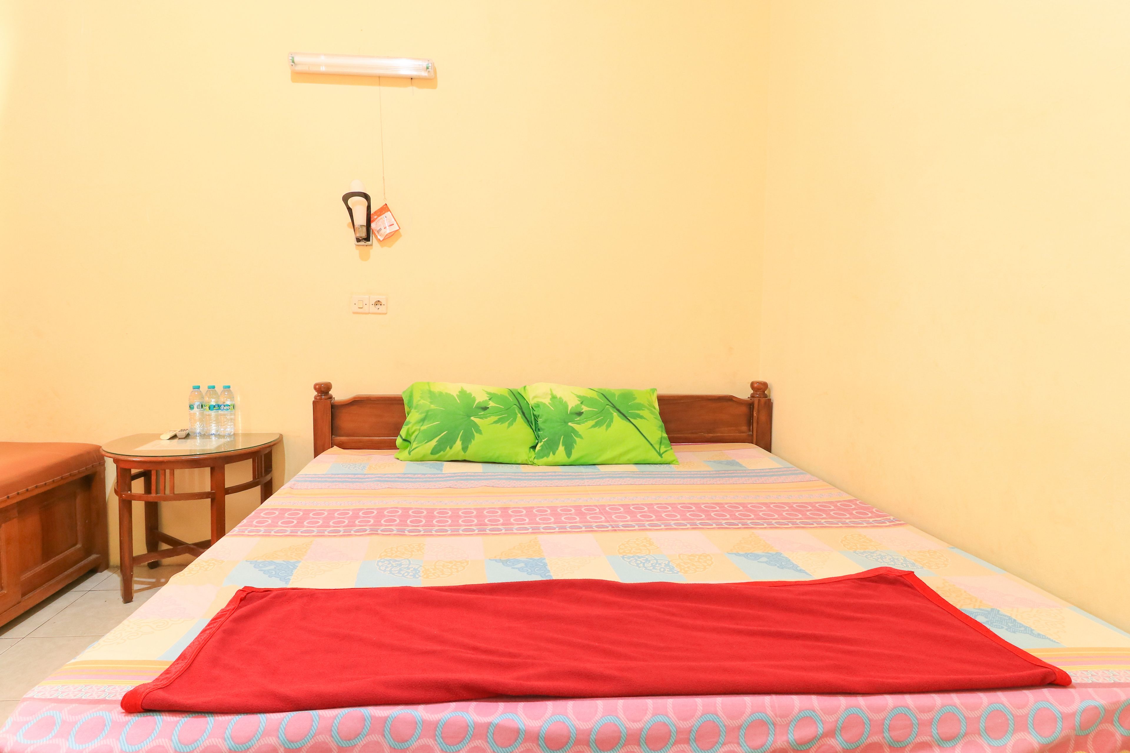 A room with a bed, a small table with water bottles, a green pillow, and a red blanket.