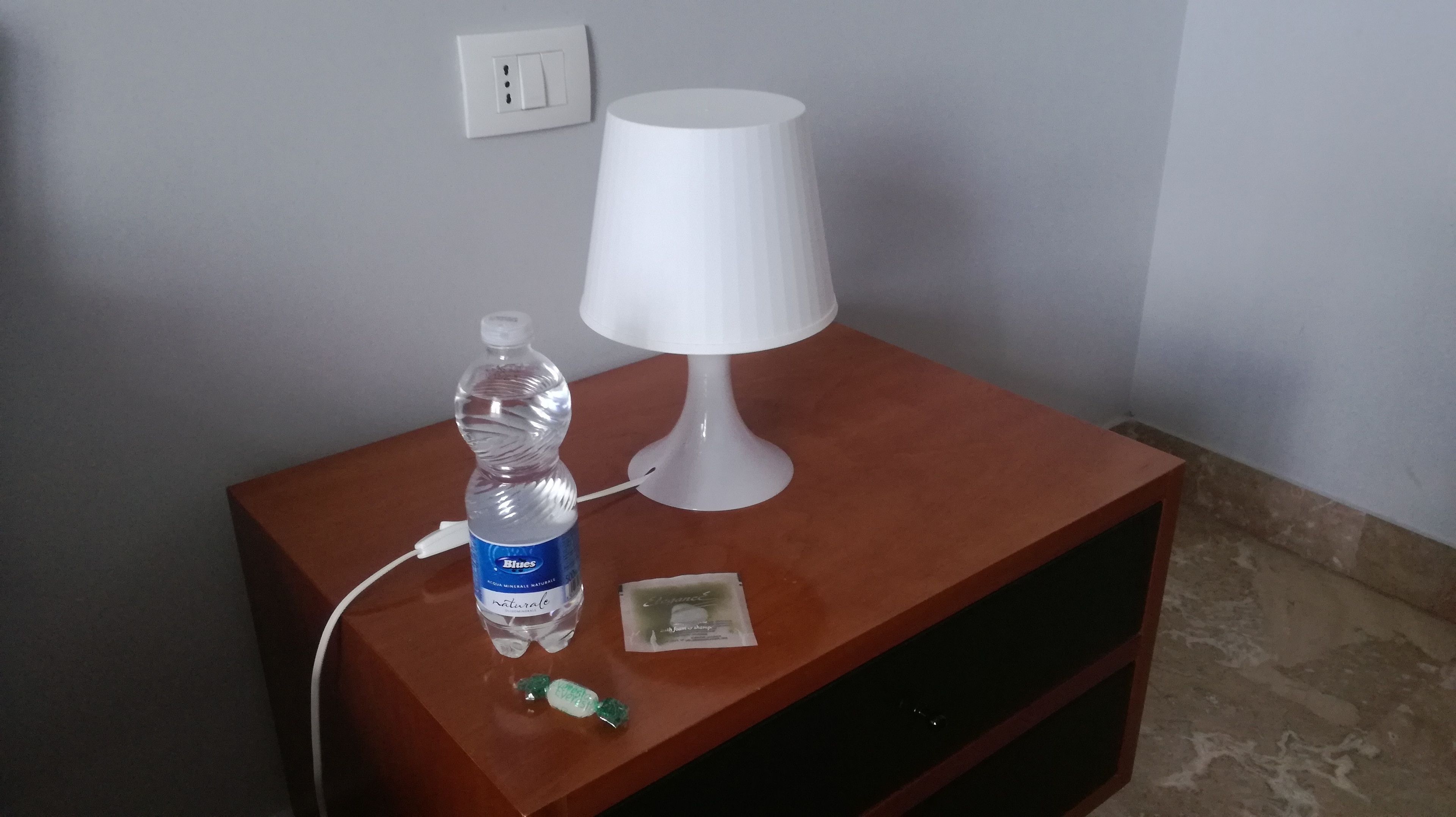 A small wooden nightstand with a white lamp, a water bottle, and a packet of gum.