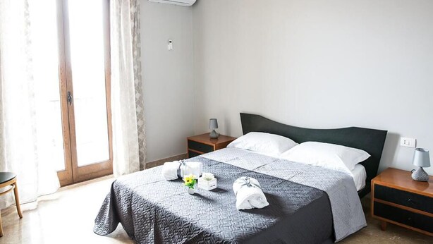 A hotel room with a bed, two bedside tables, a nightstand, a window with curtains, and an air conditioning unit. - Home La Kore (Agrigento)