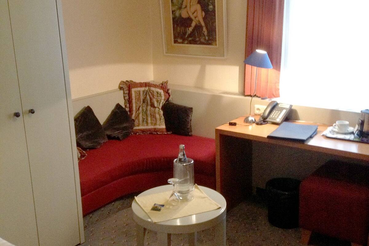 A hotel room with a bed, a desk with a lamp, a small table with a water bottle, a red sofa, and a painting on the wall.
