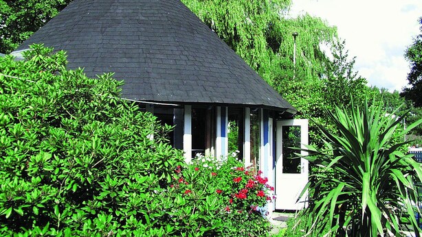 A small, single-story building with a dark roof, surrounded by lush greenery and flowering plants. - Haus am Meer (Wunstorf)