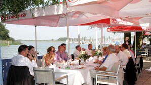 A group of people dining at an outdoor table with a view of the water and boats in the background. - Haus am Meer (Wunstorf)