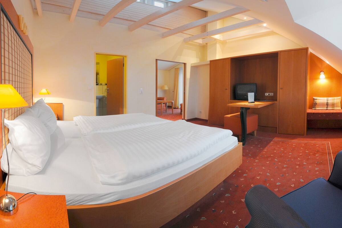 A hotel room with a large bed, a desk with a computer, a chair, and a small table with a lamp.