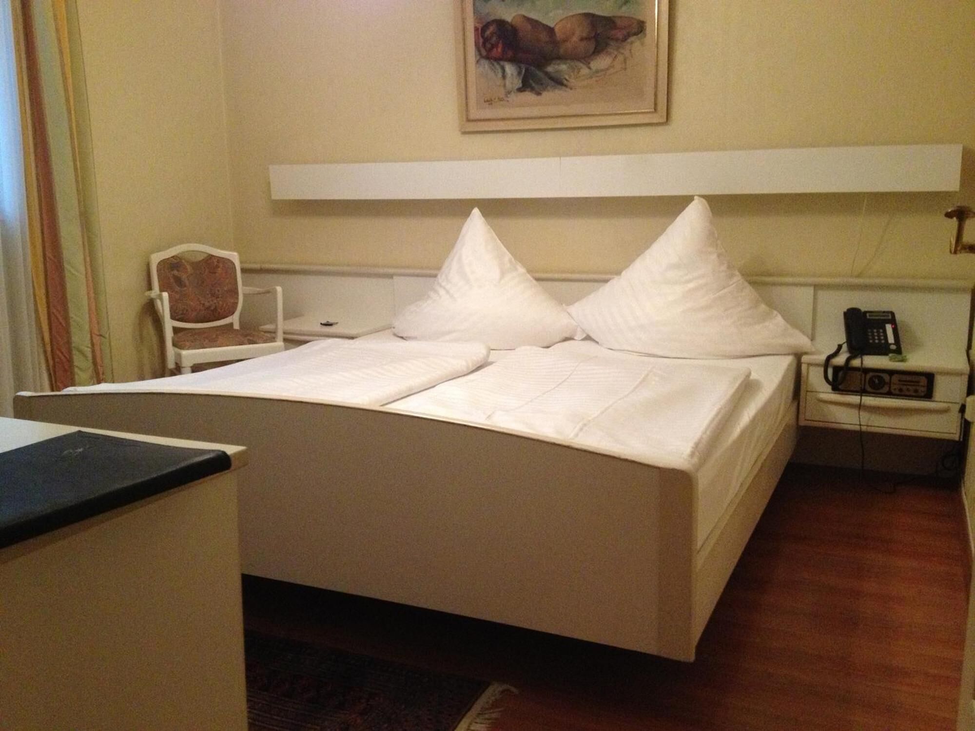 Quarto de hotel com cama, cadeira, mesa de cabeceira, telefone e um quadro na parede.