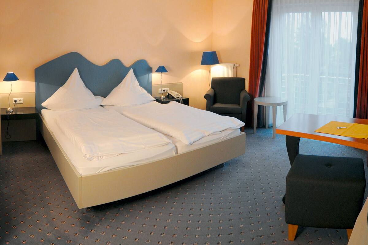 A hotel room with a large bed, two bedside tables with lamps, a chair, a small round table, and a window with curtains.