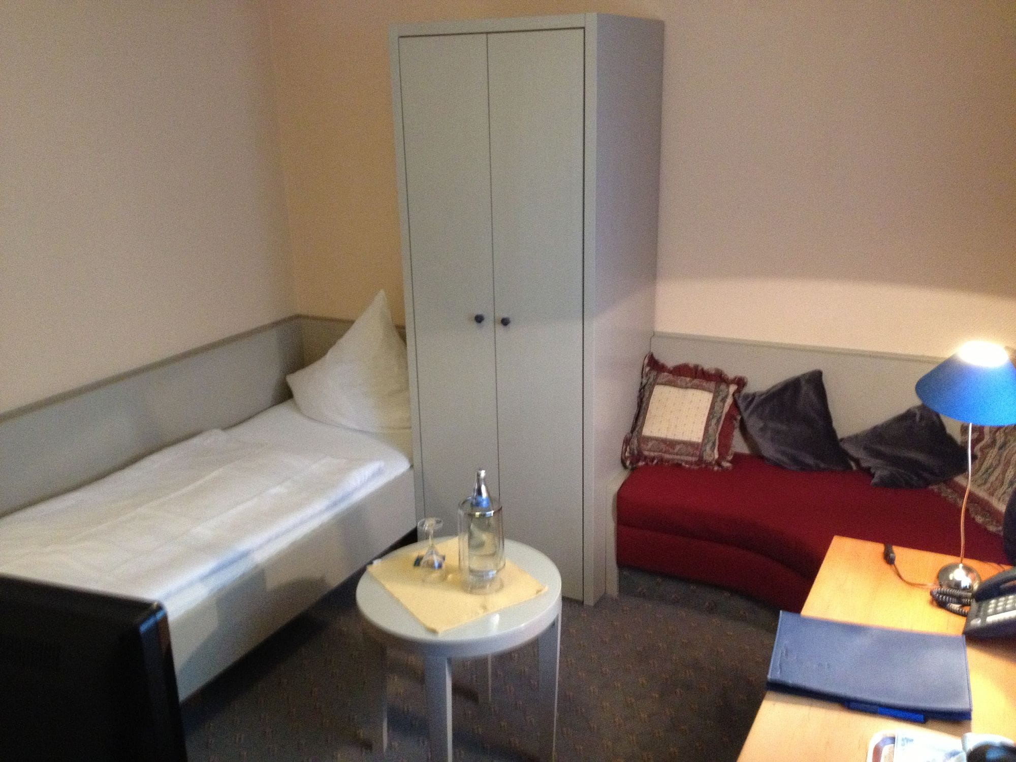 Een kleine hotelkamer met een enkel bed, een kledingkast, een klein tafeltje met een lamp en een bureau met telefoon en notitieboekje.