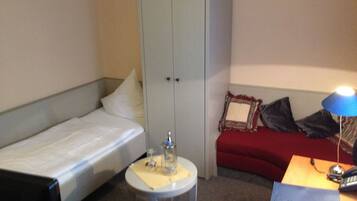 Een kleine hotelkamer met een enkel bed, een kledingkast, een klein tafeltje met een lamp en een bureau met telefoon en notitieboekje.