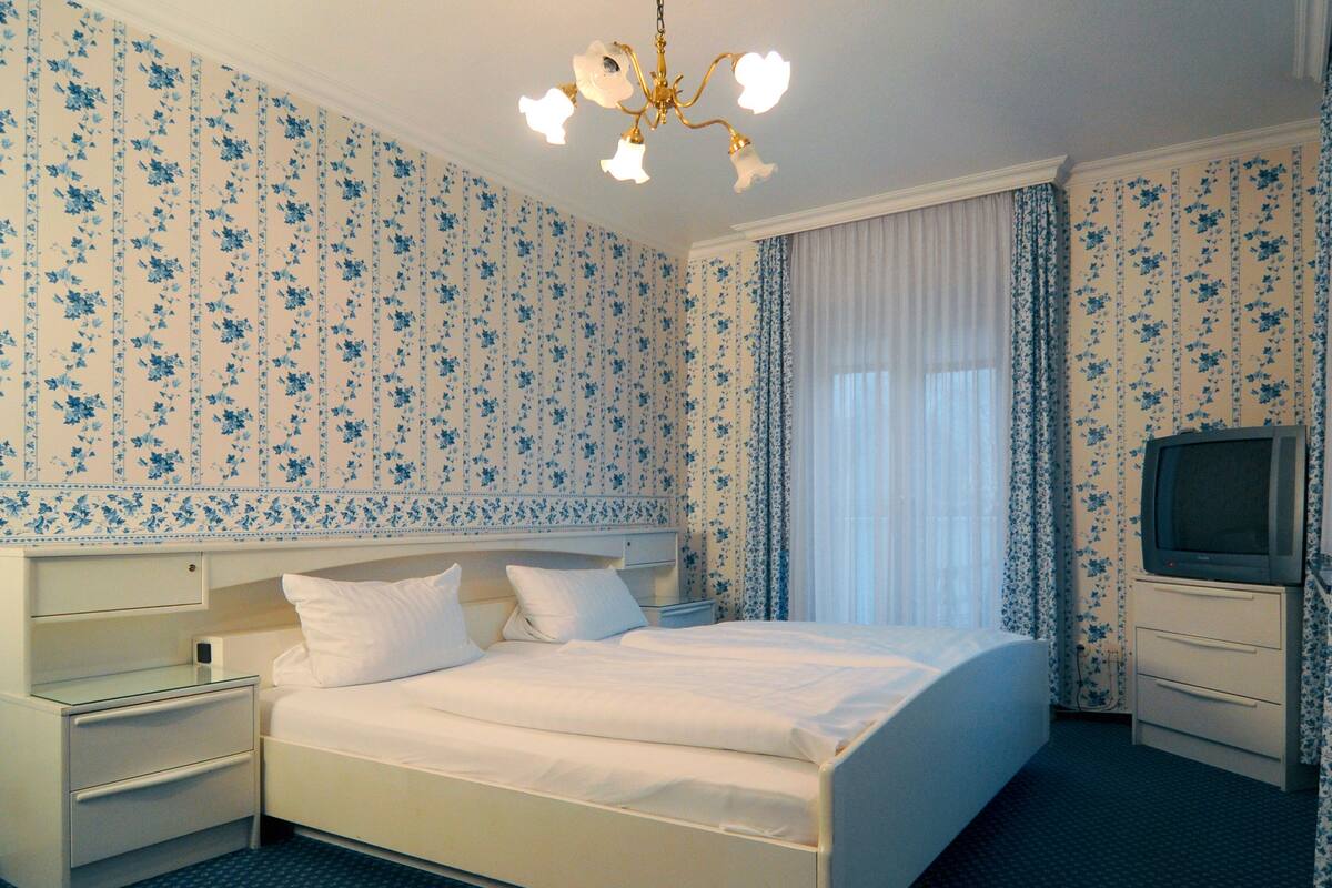 A hotel room with a large bed, a bedside table, a TV, and floral wallpaper.