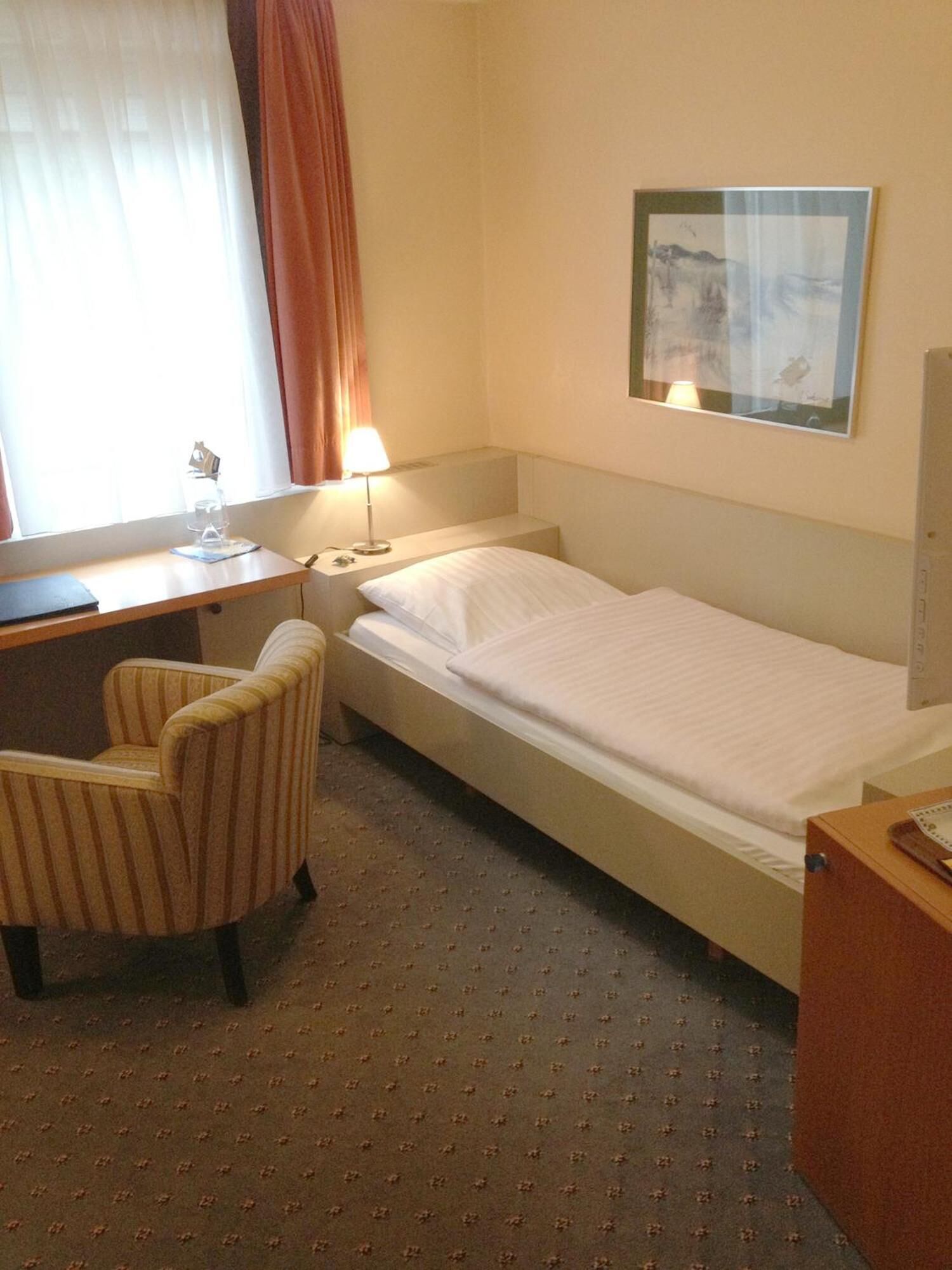 A hotel room with a single bed, a desk with a chair, a lamp, and a painting on the wall.