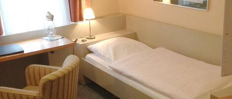 Habitación de hotel con una cama individual, un escritorio con silla, una lámpara y un cuadro en la pared.