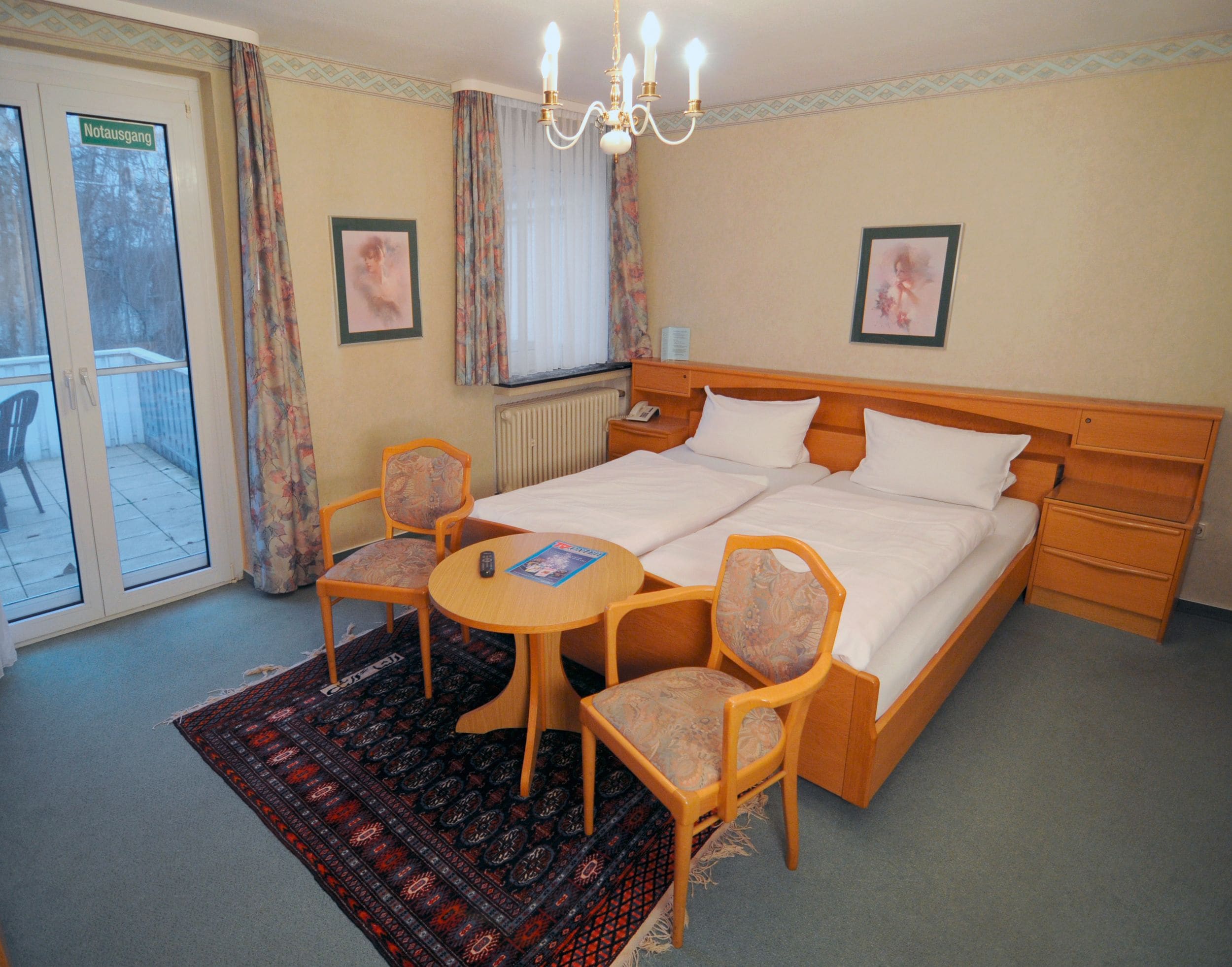 A double bed with a wooden headboard, two bedside tables, a small round table with two chairs, a patterned rug, and a view of a balcony with a door labeled "Notausgang".
