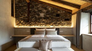 A bed with a stone wall behind it, wooden beams on the ceiling, and a window with curtains.