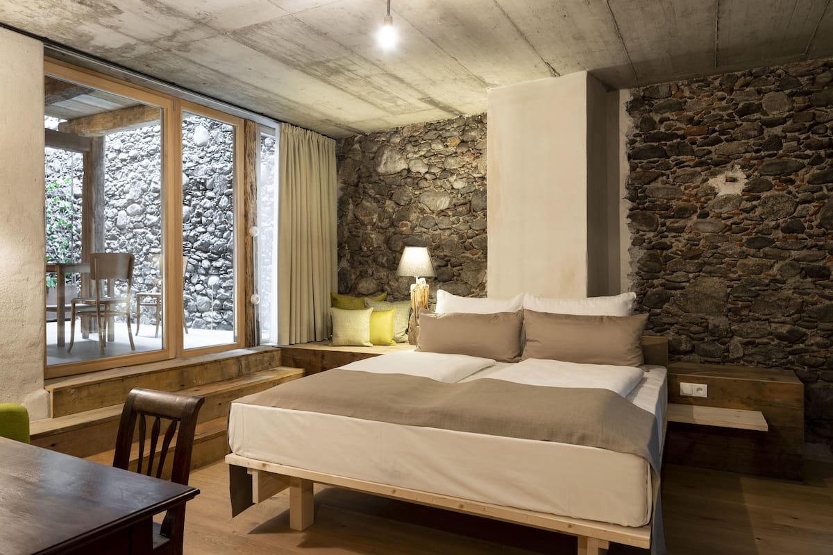 Quarto com parede de pedra, cama de madeira, mesa de cabeceira, luminária e janela com cortinas.