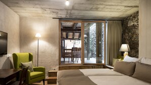A bedroom with a bed, a green armchair, a desk with a chair, a lamp, and a view of an outdoor area with a table and chairs.