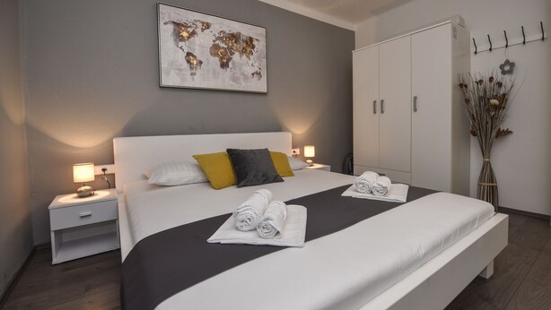 A neatly made bed with white and gray linens, a world map artwork above, a white wardrobe, and a small nightstand with a lamp. - Holiday home Klarisa (Novalja)