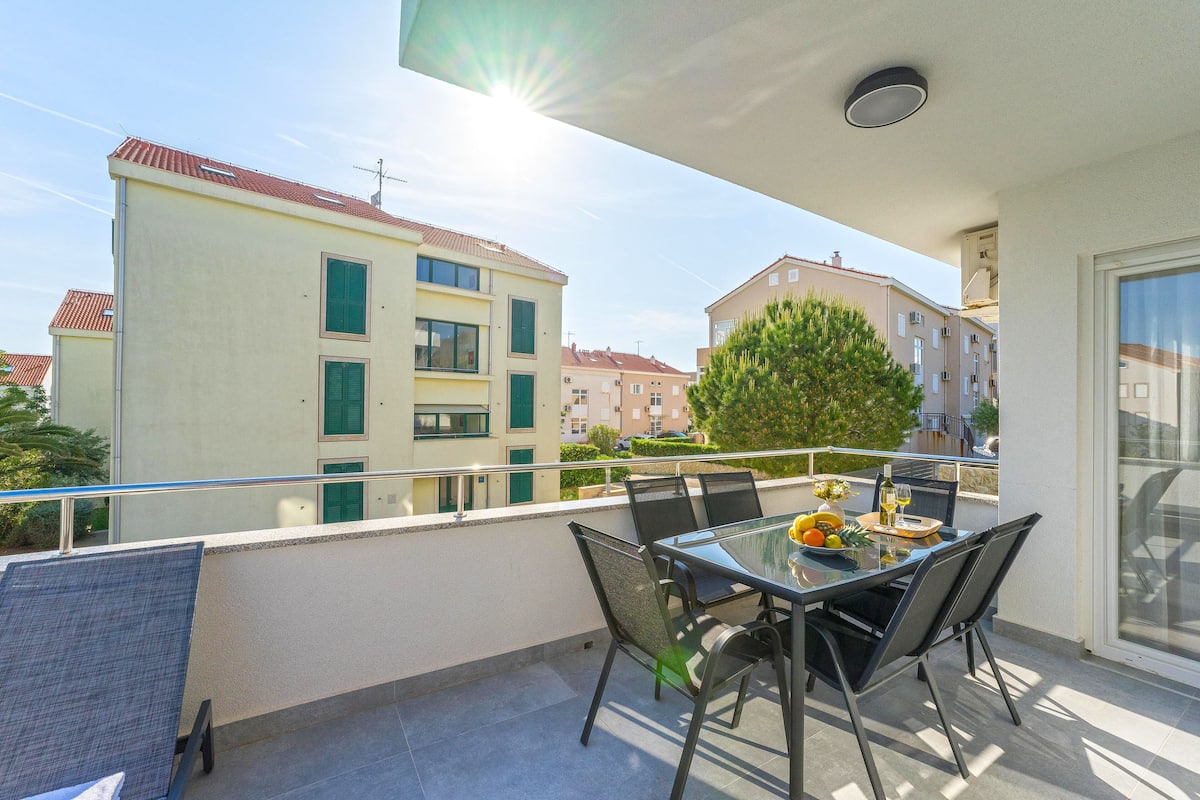 Un balcon avec une table et des chaises en verre, donnant sur un quartier résidentiel avec des immeubles et un arbre.