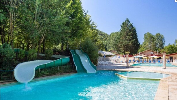A swimming pool with two slides, surrounded by trees and a poolside area with lounge chairs.