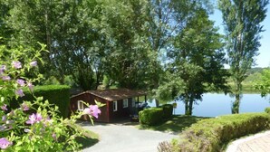 A small cabin with a gravel driveway leading to a lake, surrounded by lush greenery and trees.
