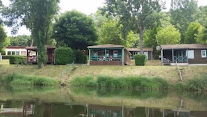 A row of cabins by a lake with a grassy area in front and trees in the background.