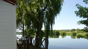 A wooden deck with steps leading to a white building, overlooking a calm lake with trees and hills in the background.