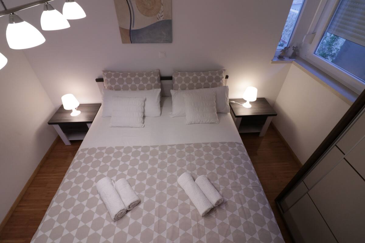 A neatly arranged bedroom with a patterned bedspread, two bedside lamps, and a window with a view.
