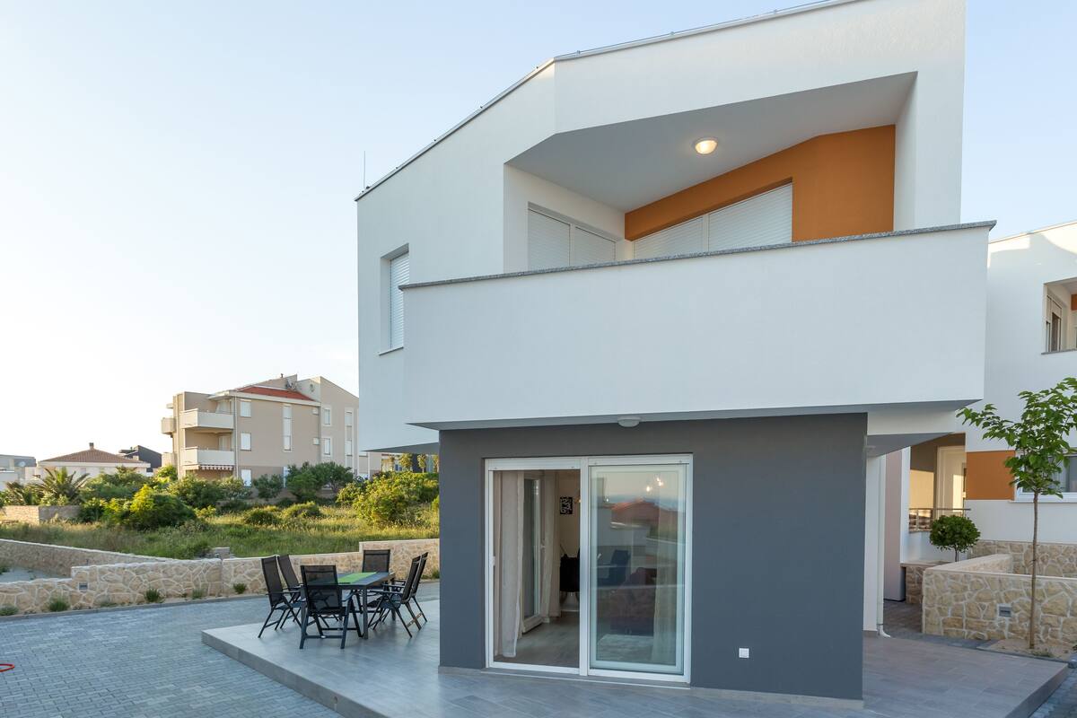A modern two-story building with a balcony, a patio with outdoor furniture, and a stone wall.