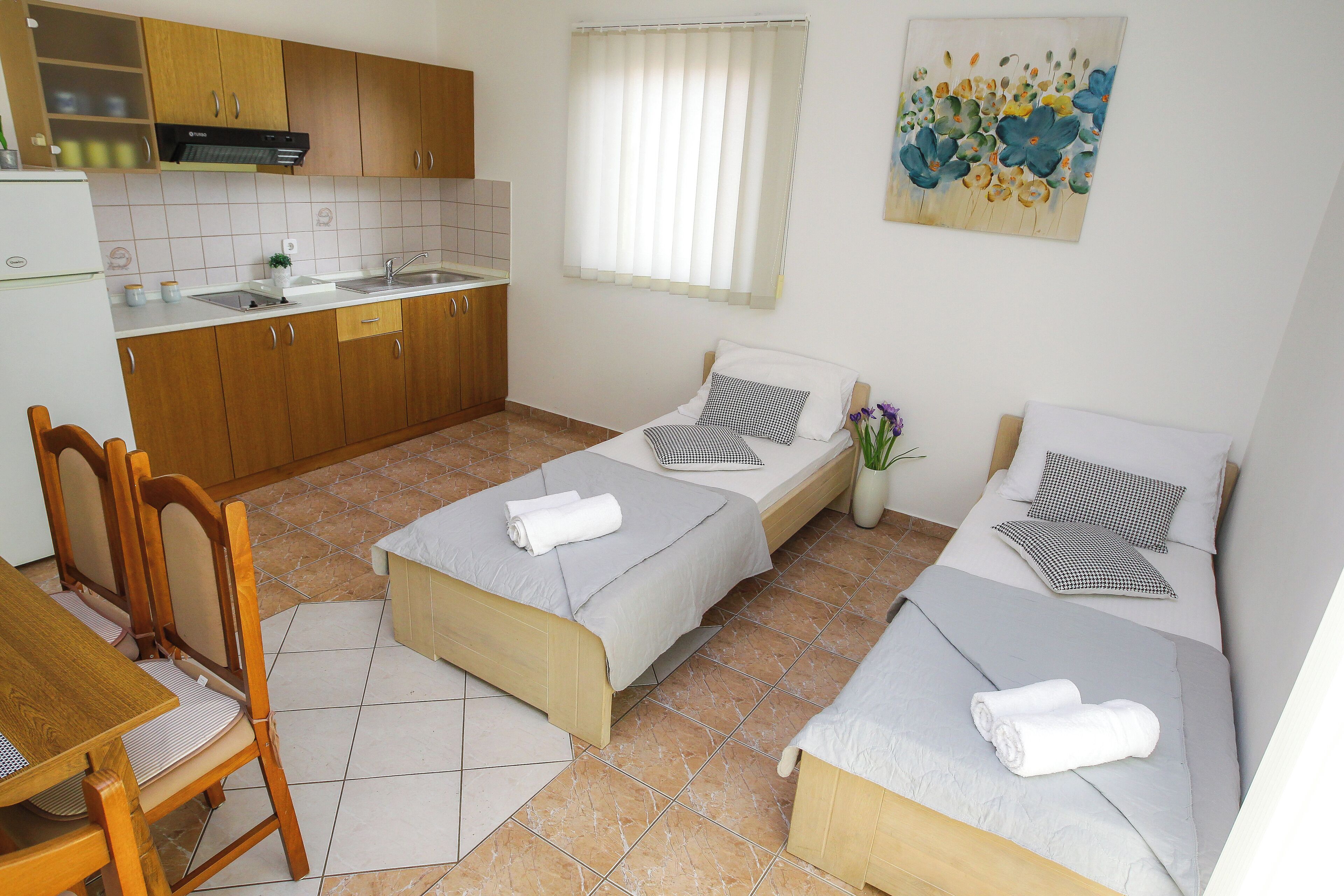 A compact hotel room with two single beds, a small kitchenette, a dining area with a table and chairs, and a bathroom with a shower.