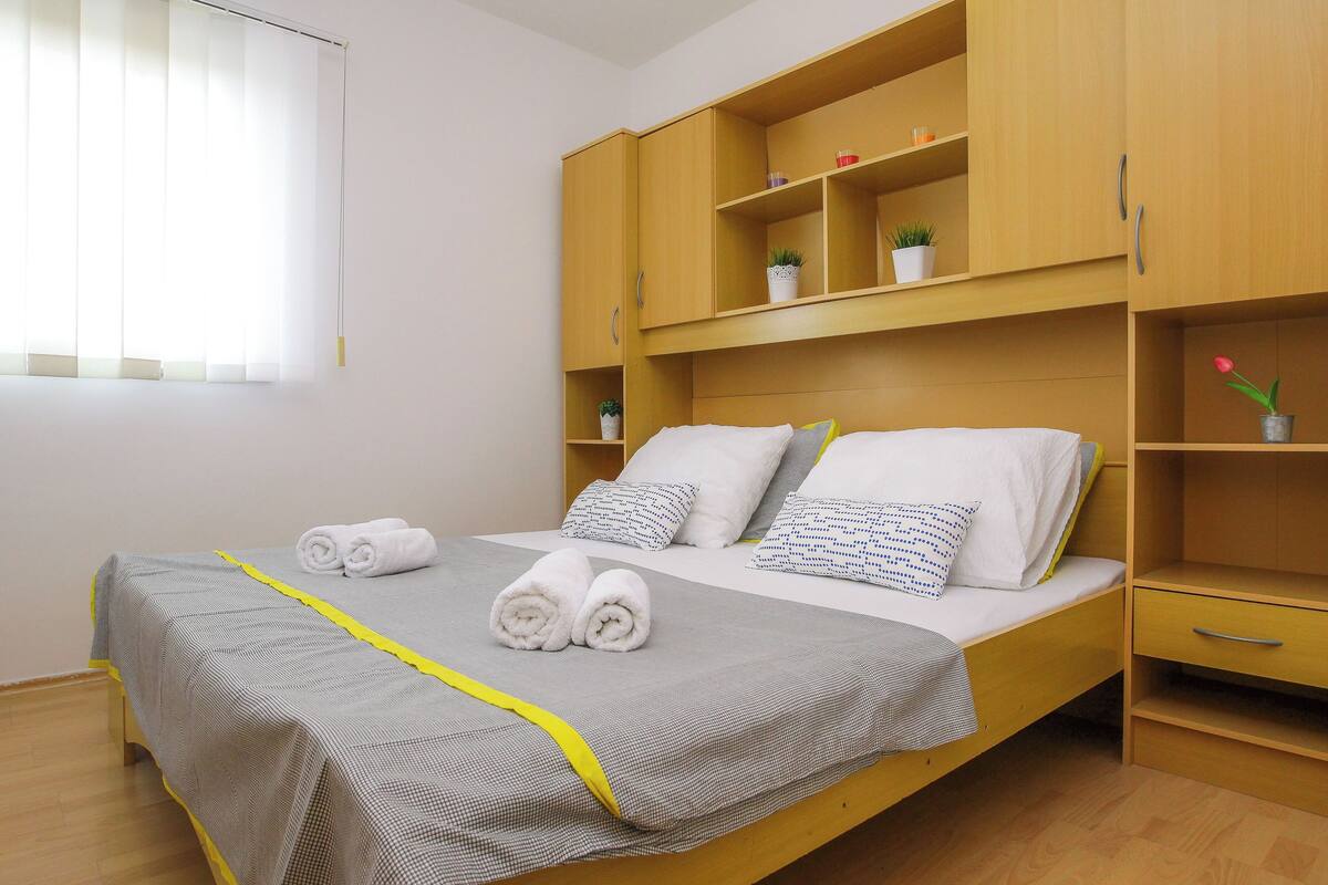 A neatly made bed with rolled towels, a bedside table, and a wooden wardrobe with shelves.