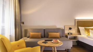 A hotel room with a sofa, a bed, a small table with a plate of fruit, a chair, and a bedside table with a lamp.