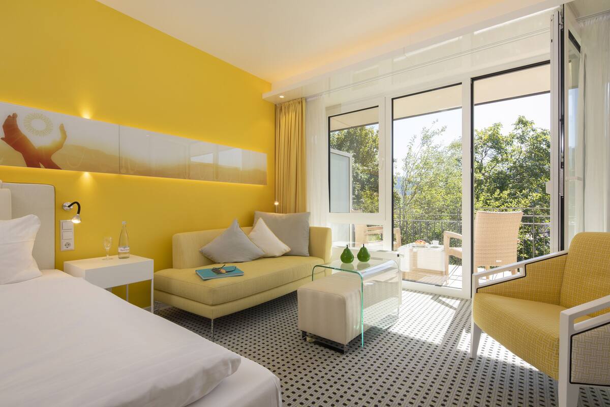 Ein Hotelzimmer mit einem Bett, einem Sofa, einem kleinen Tisch und einem Sessel. Ein großes Fenster bietet Blick auf Bäume.
