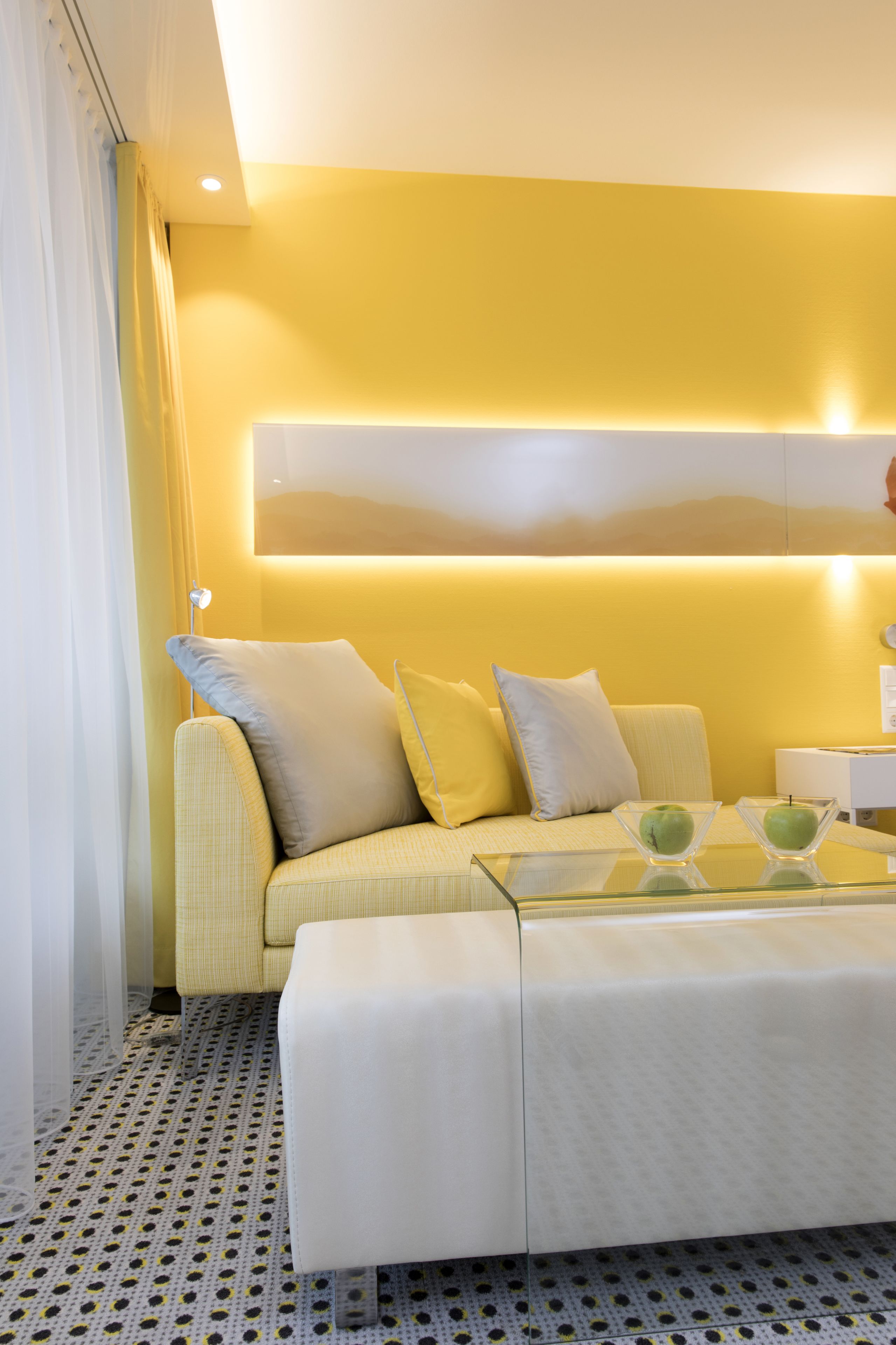Een moderne woonkamer met een geel accentmuur, een bank met kussens, een glazen bijzettafeltje met appels en een vloer met een patroon.