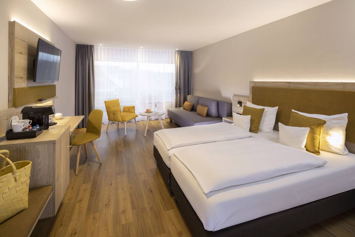 Ein Hotelzimmer mit einem Bett, einem Schreibtisch mit Kaffeemaschine, einem Sessel, einer Couch und einem an der Wand befestigten Fernseher.