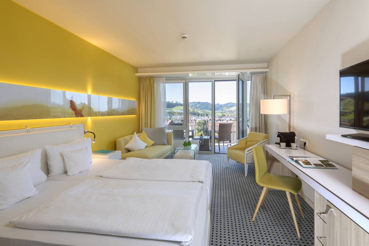 Ein Hotelzimmer mit einem großen Bett, einem Schreibtisch mit Stuhl, einer Sitzecke und Blick auf die Stadt durch große Fenster.