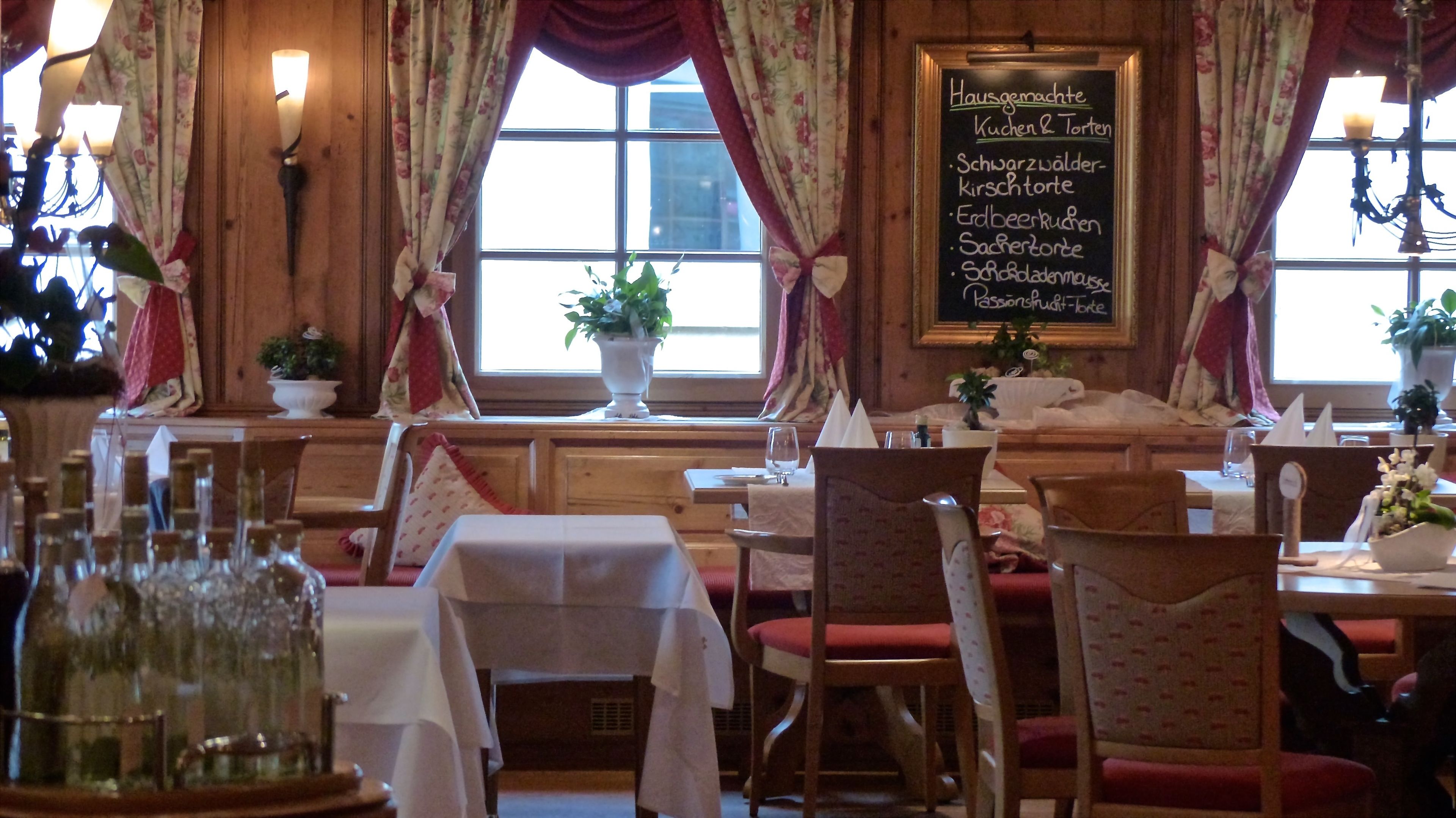 L’intérieur d’un restaurant, avec des meubles en bois, des rideaux à motifs floraux et un tableau noir affichant le menu et les plats proposés.