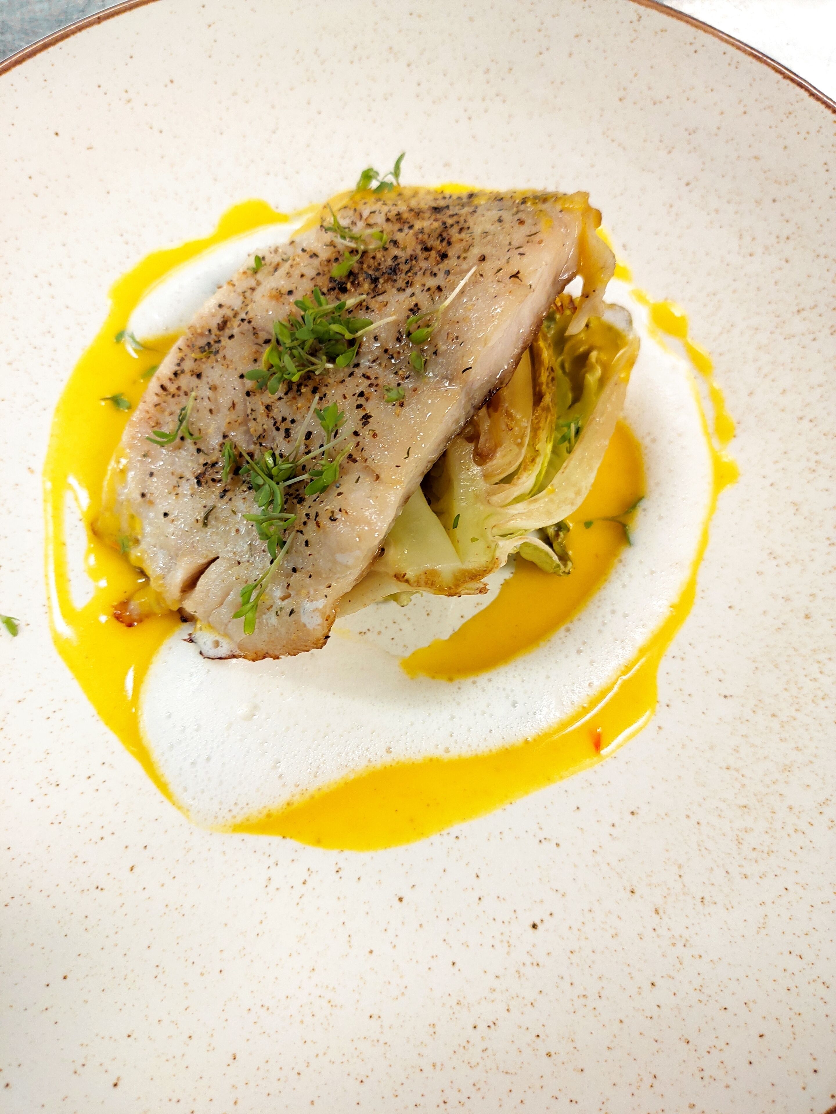 A plate with a piece of fish, greens, and a yellow sauce.