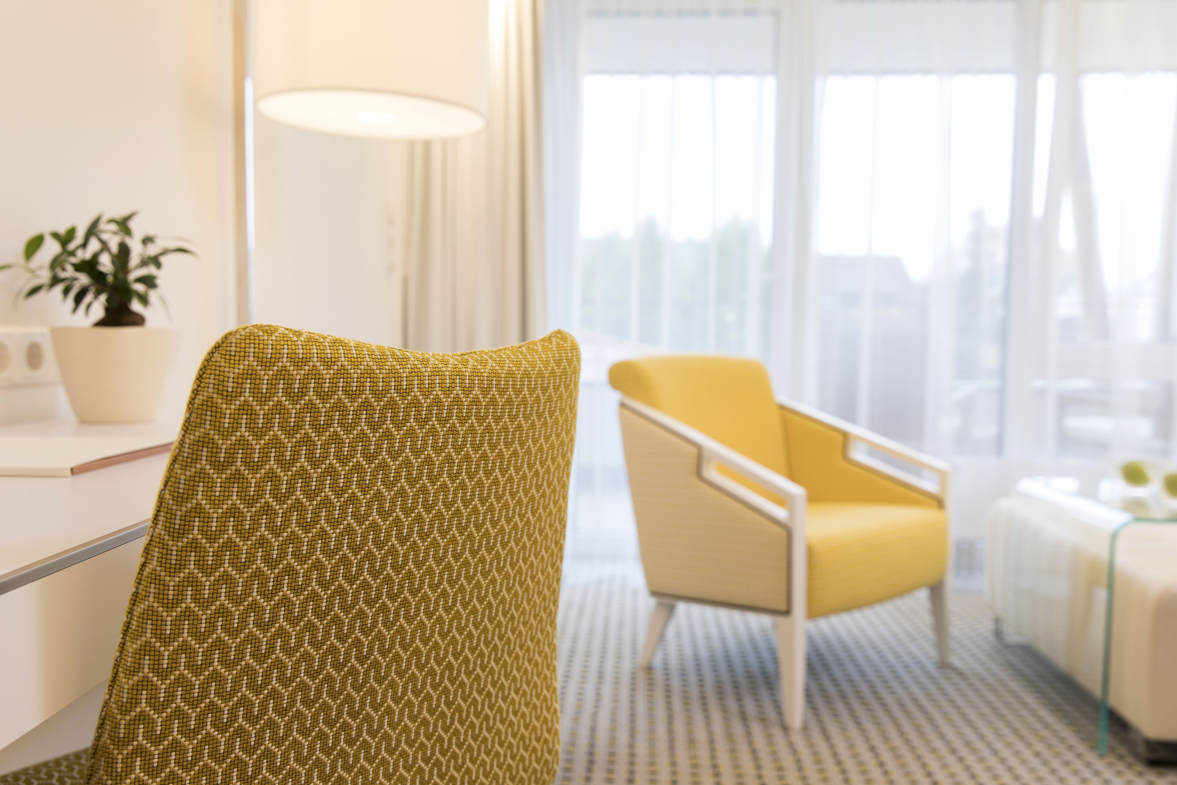 A modern hotel room with a patterned armchair, a yellow armchair, a bedside table, and a window with a city view.