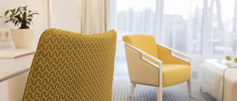 A modern hotel room with a patterned armchair, a yellow armchair, a bedside table, and a window with a city view.