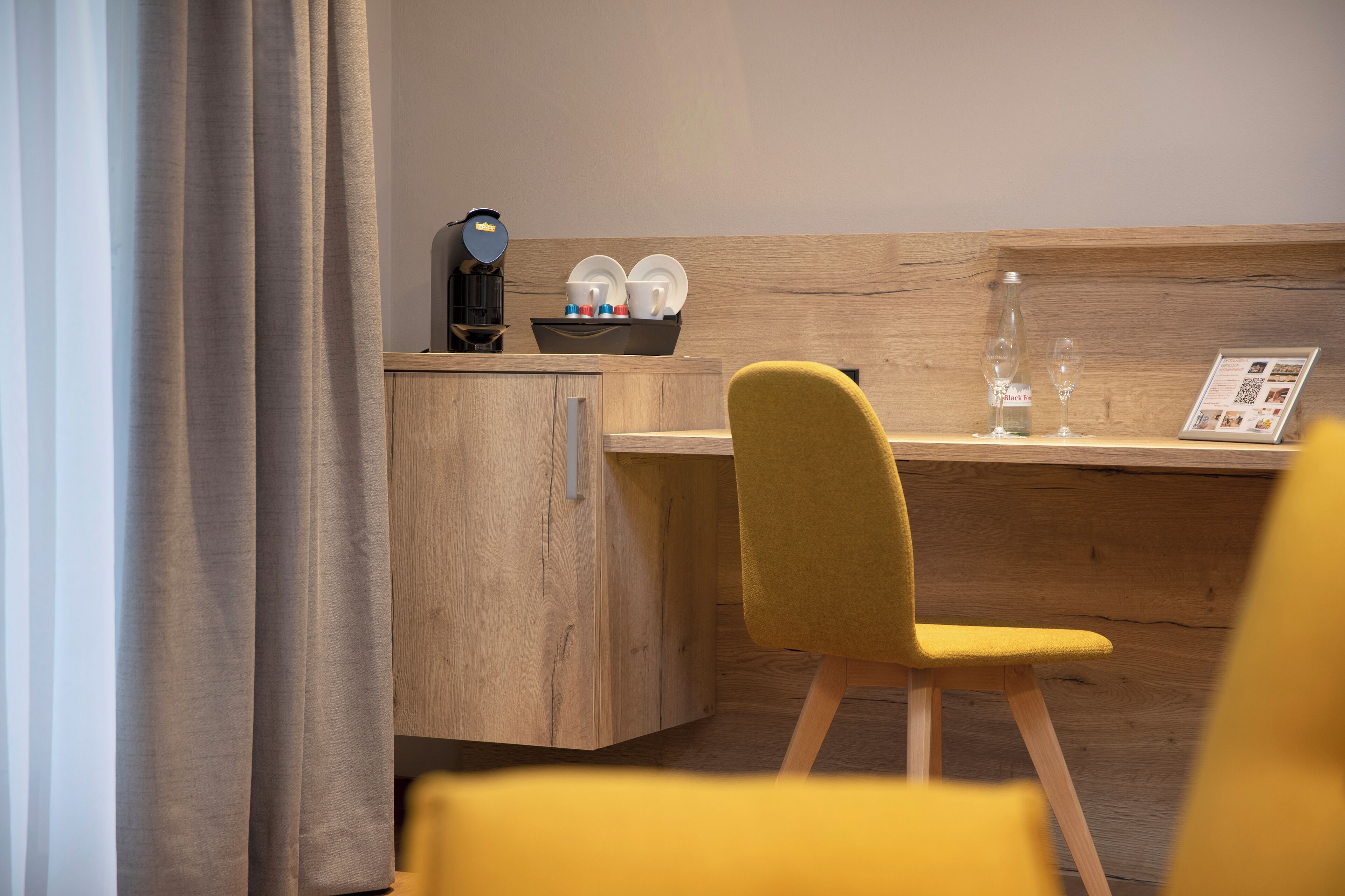 Un bureau en bois, une machine à café, un plateau avec des tasses et une bouteille, et une chaise avec un coussin jaune.