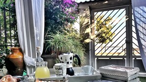 A table set with breakfast items, including pastries, a drink, and a coffee pot, in an outdoor patio area with plants and a view of a building. - Il Ritrovo di Ulisse Bed & Breakfast (Villa San Giovanni)