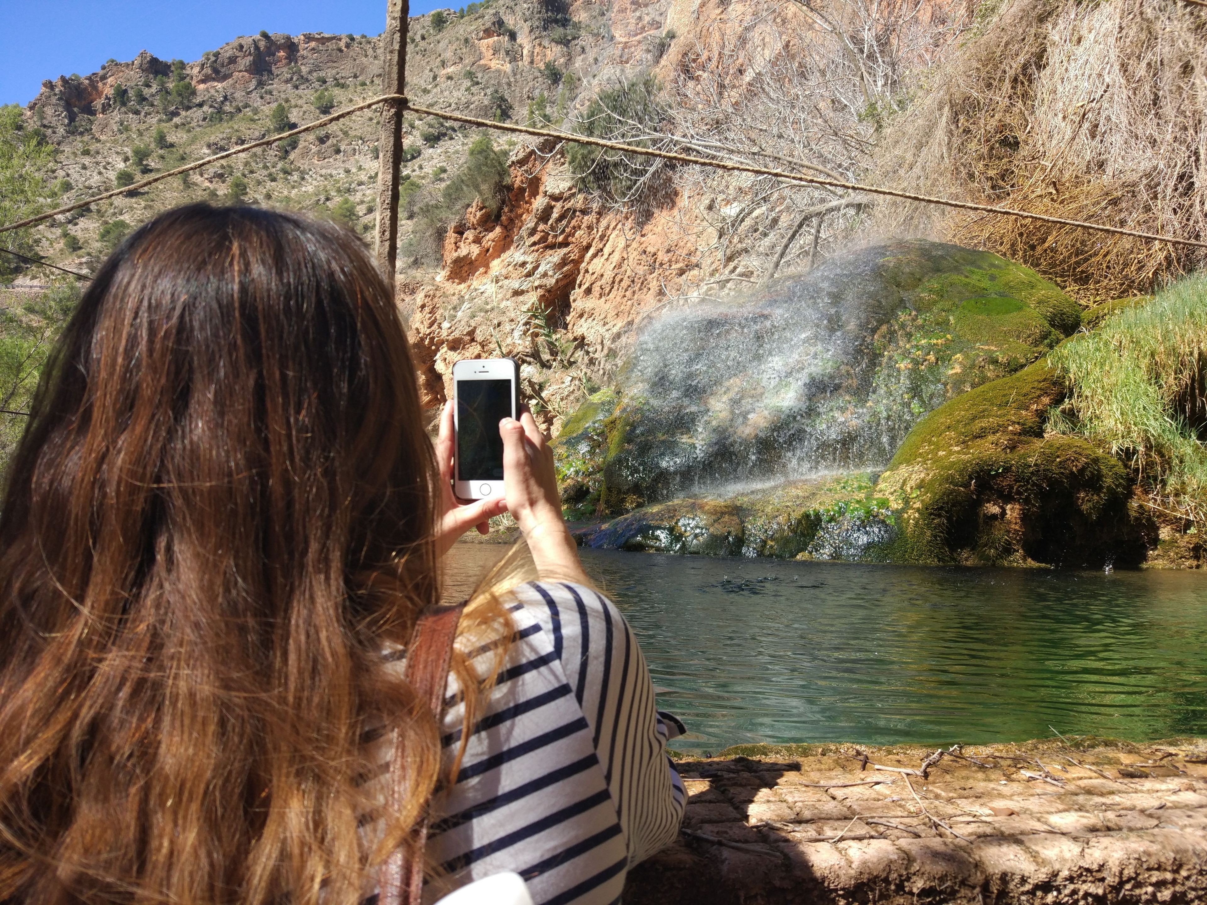Una persona fotografiando un paisaje natural con una masa de agua, terreno rocoso y vegetación.
