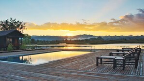 A wooden deck with a pool, lounge chairs, and a view of mountains at sunset.
