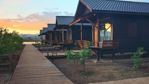 A row of wooden cabins with a wooden deck leading to them, surrounded by small plants and a view of the mountains in the background.