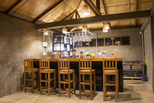 A bar area with wooden chairs, a bar counter, hanging light fixtures, and a window with blinds.