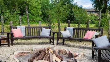 A wooden bench with cushions, a fire pit, and a gravel ground surrounded by trees.
