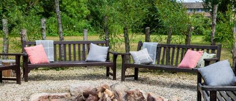 A wooden bench with cushions, a fire pit, and a gravel ground surrounded by trees.