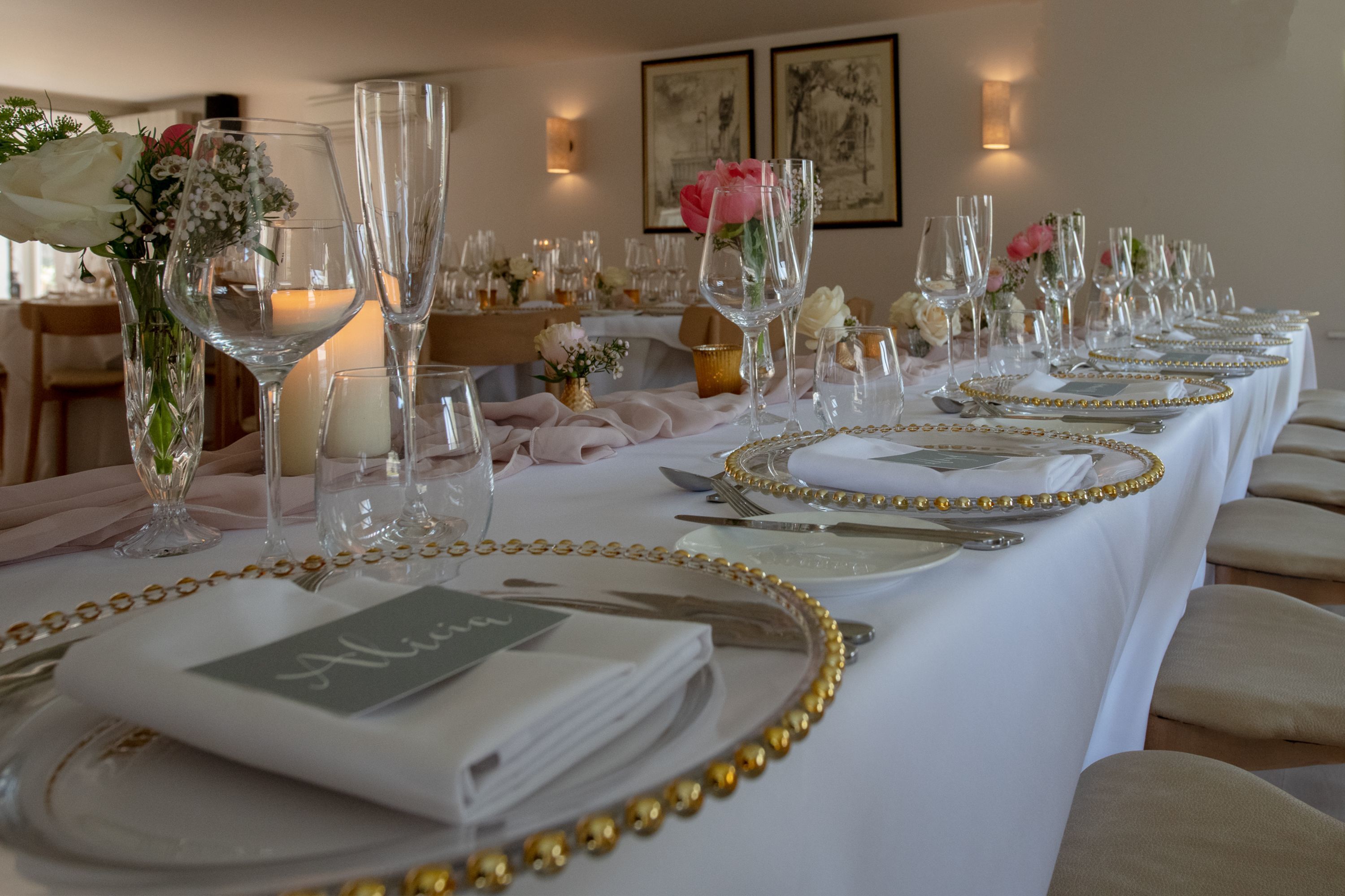 A long dining table set with gold-rimmed plates, clear glassware, and white napkins with a name card.