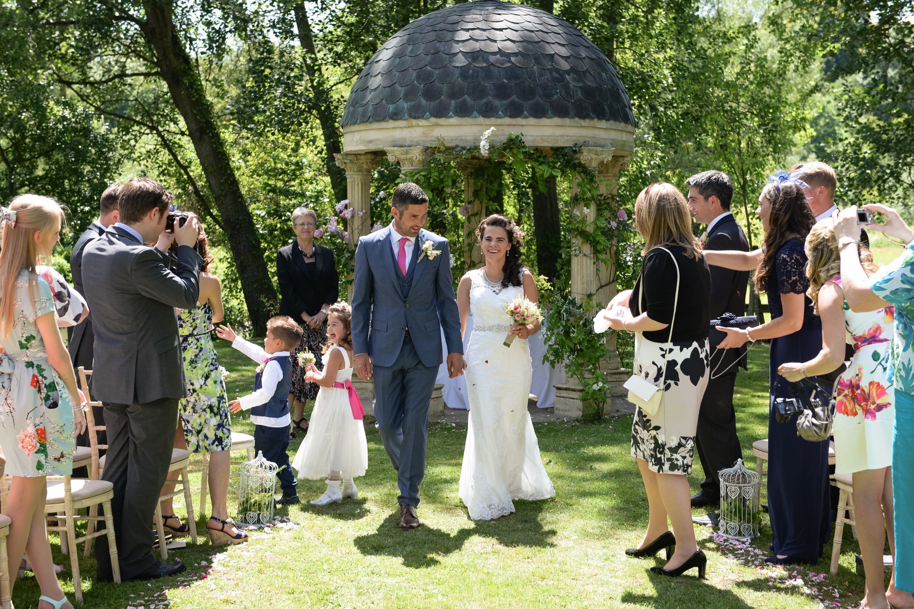 Une cérémonie de mariage dans un jardin, avec un kiosque, des invités, ainsi qu’une mariée et un marié.